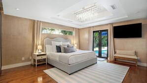 A modern bedroom with a large bed, a chandelier, a TV, and a view of a pool. - Pool/Spa & Walk to the Beach Ping Pong Billiards (Palm Beach Shores)