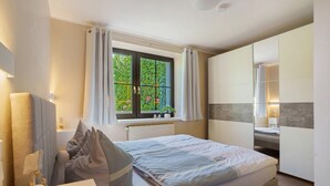 Et soveværelse med seng, sengebord, et vindue med gardiner, en radiator og udsigt til grønne områder.