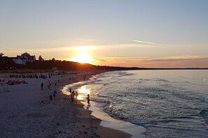 Una spiaggia con persone che camminano lungo la riva, un tramonto sullo sfondo e edifici in lontananza.
