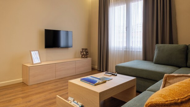 A modern living room with a flat-screen TV mounted on the wall, a wooden coffee table with books, a green sofa, and a wooden cabinet.