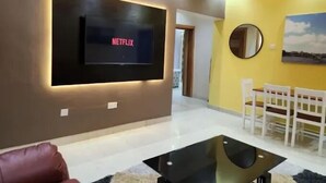 Moderni olohuone, jossa on litteänäyttöinen televisio, jossa näkyy teksti »NETFLIX», valkoiset tuolit ruokailutilassa sekä keltainen tehosteseinä.