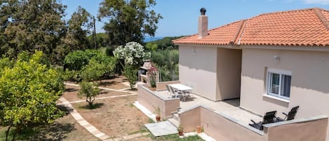A house with a tiled roof, a patio with chairs, and a garden with trees and shrubs.