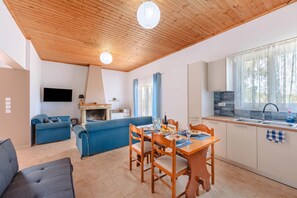 A modern living room with a blue sofa, a dining table with chairs, a kitchen area with a sink and stove, and a TV mounted on the wall. - To Pournari Beachside Garden House (Pyrgos)