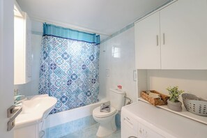 Un baño con cortina de ducha estampada, un lavabo blanco, un inodoro y un mueble con cestas y una planta en maceta.