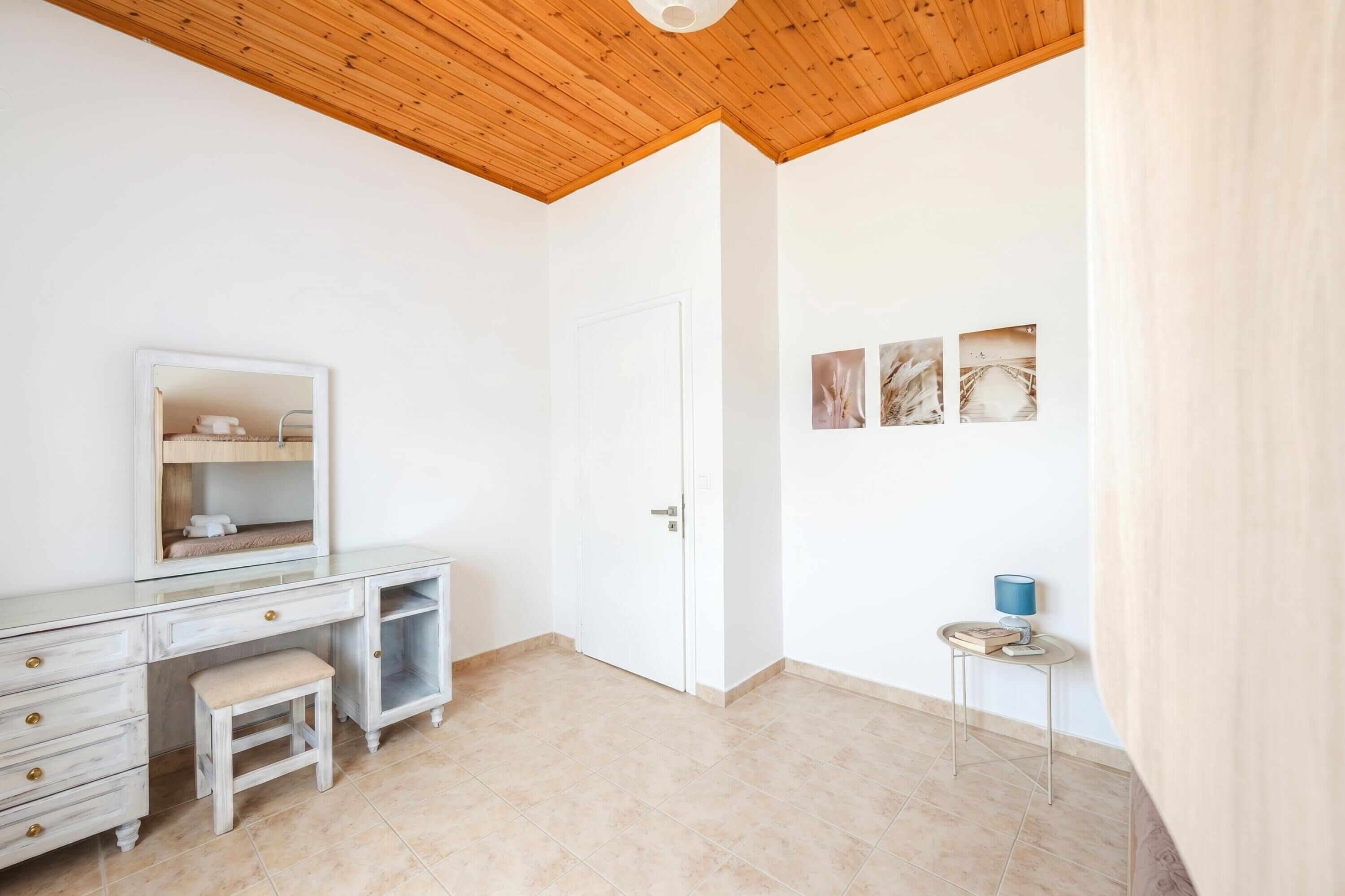 Une pièce avec un plafond en bois, une porte blanche, une coiffeuse avec un miroir, un petit tabouret et une table d’appoint avec une lampe.