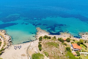An aerial view of a coastal area with clear blue waters, rocky terrain, and a few buildings with visible structures.