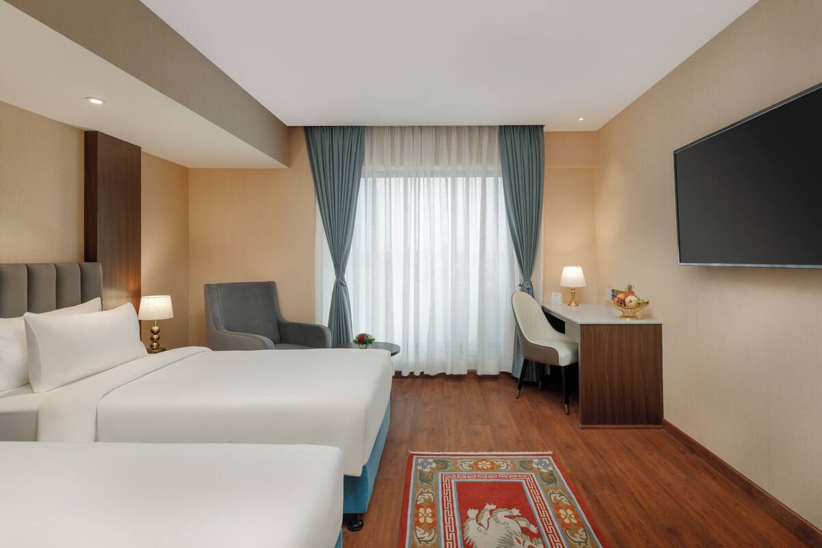 A hotel room with two beds, a desk, a chair, a TV, and a window with curtains.
