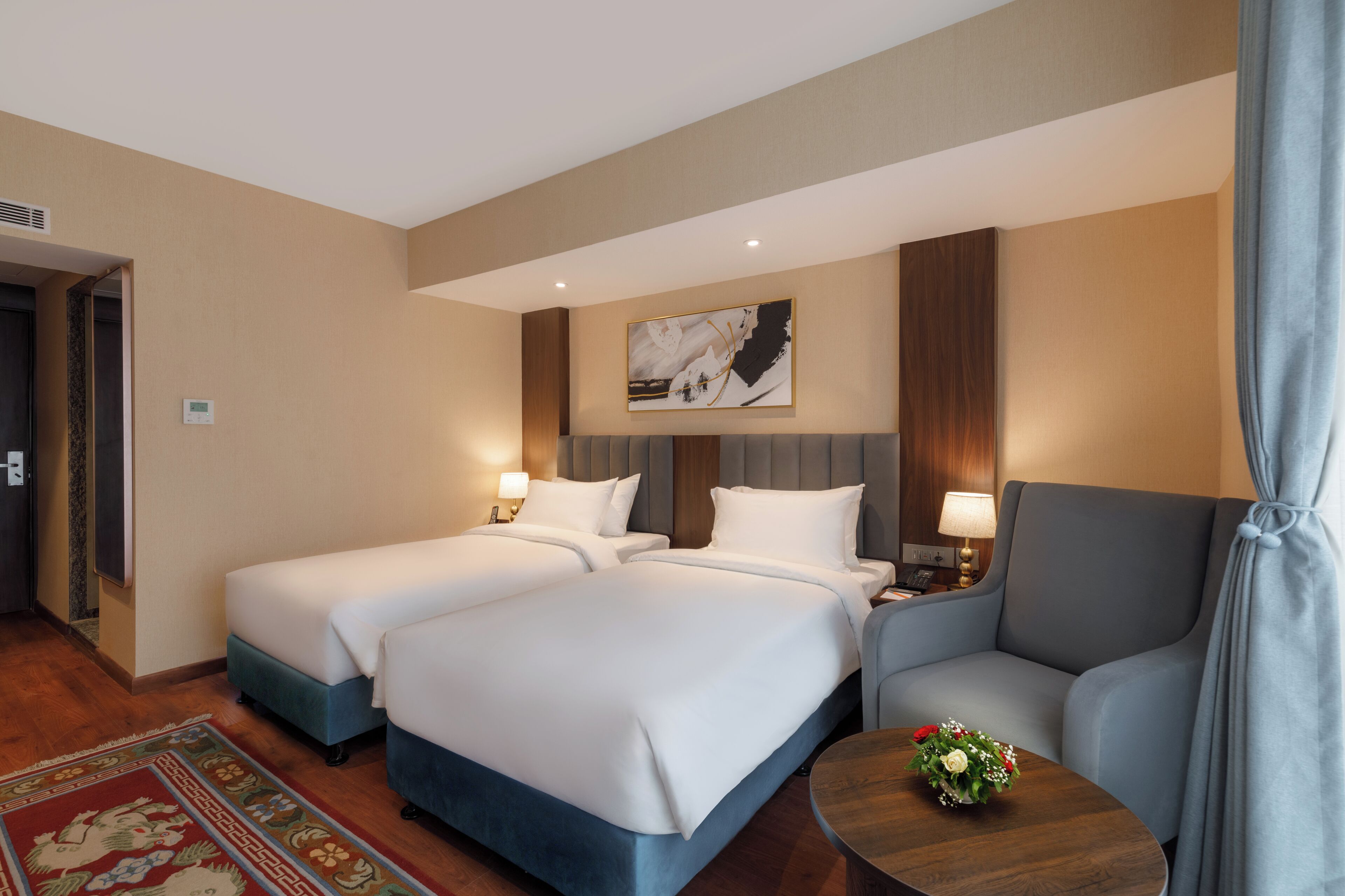 A hotel room with two beds, a sofa, a small table with a vase, and a painting on the wall.