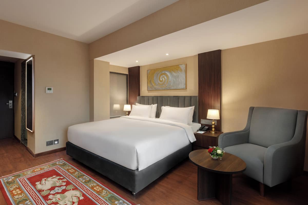 A hotel room with a large bed, a grey armchair, a small table with a vase, and a painting on the wall.