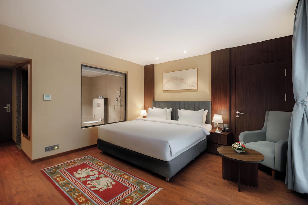 A hotel room with a large bed, a bedside table with a lamp, a chair, a wooden wardrobe, and a mirror.