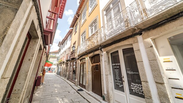 A narrow street with buildings on either side, featuring balconies with metal railings and shop signs.