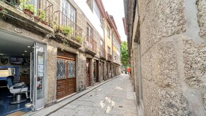A narrow street with buildings on either side, a visible shop on the left, and a stone wall on the right.