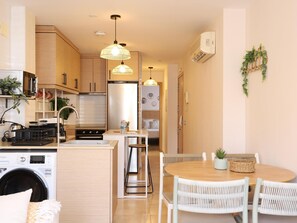 A modern kitchen with a built-in washer, a dining area with a round table and wicker chairs, and pendant lighting.