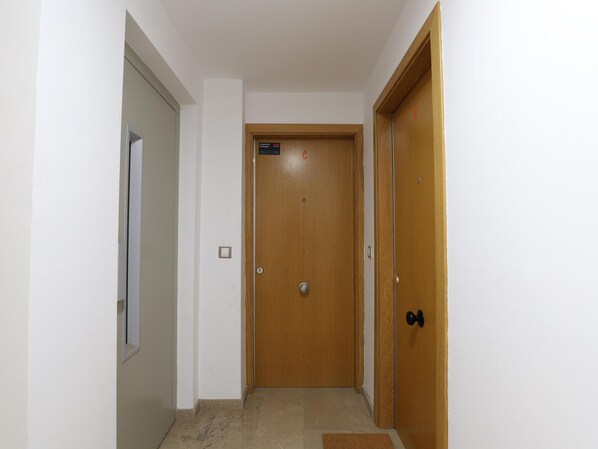 A narrow hallway with a wooden door at the end, a small window on the left, and a doormat on the floor.