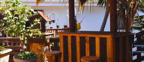 A wooden outdoor seating area with a thatched roof, surrounded by potted plants and a palm tree.