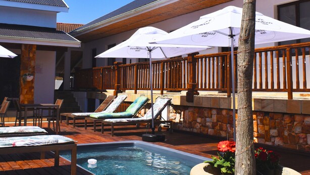 A pool area with lounge chairs and umbrellas, a wooden deck, and a stone wall. - Bergwood Inn (Groot Brakrivier)