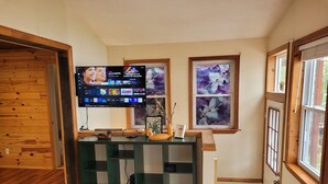 A room with a wooden floor, a TV mounted on the wall, a green entertainment unit, and two windows with artwork.