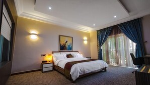 A hotel room with a bed, bedside lamps, a painting on the wall, and a view of the outdoors through the window. - Camas Hotels  (Abeokuta)