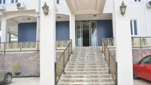 A building entrance with a staircase leading to a covered area, a brick wall, and a red car parked nearby. - Camas Hotels  (Abeokuta)
