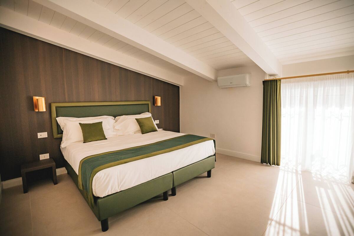 A hotel room with a large bed, a nightstand, a wall-mounted air conditioner, and a window with sheer curtains.