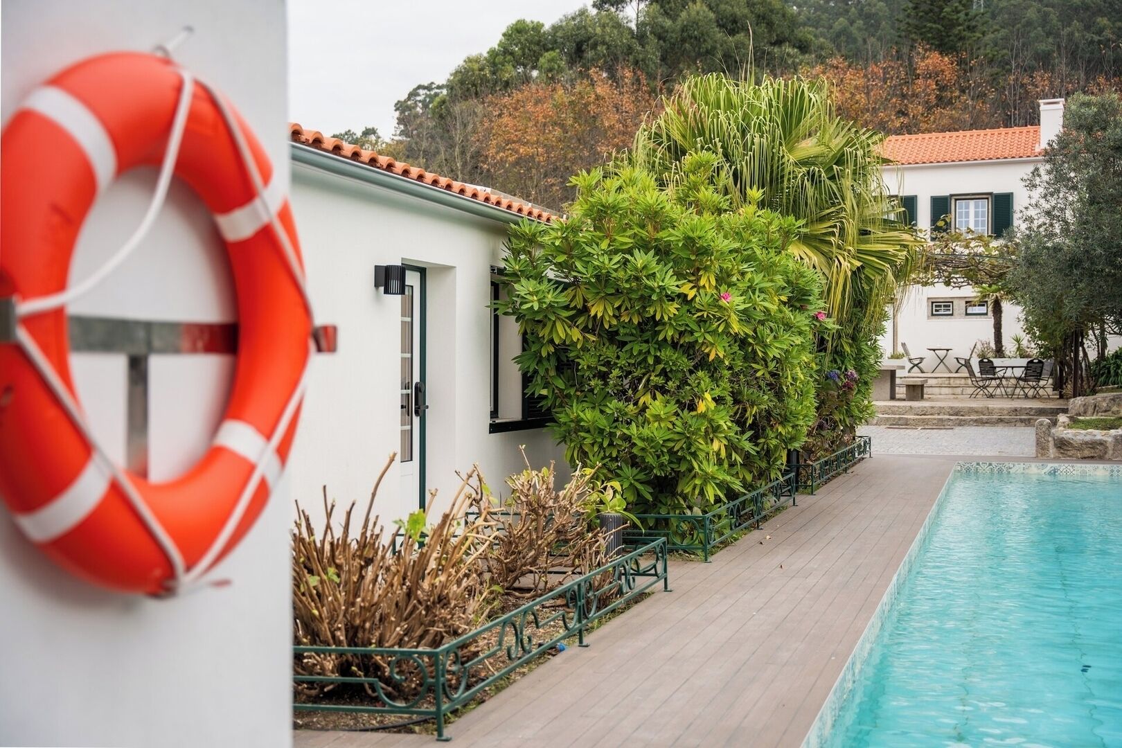 A poolside area with a lifebuoy, a white building with a tiled roof, and lush greenery.