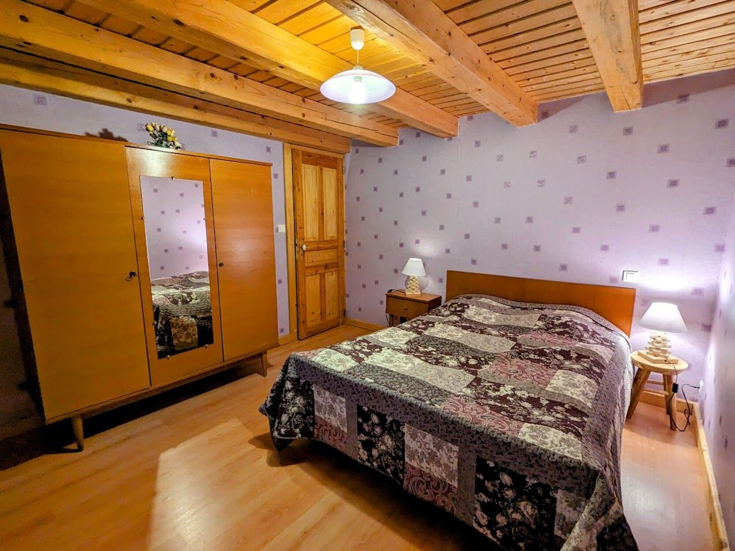 Een slaapkamer met een houten plafond, een bed met een bloemenquilt, een houten garderobe en een nachtkastje met een lamp.