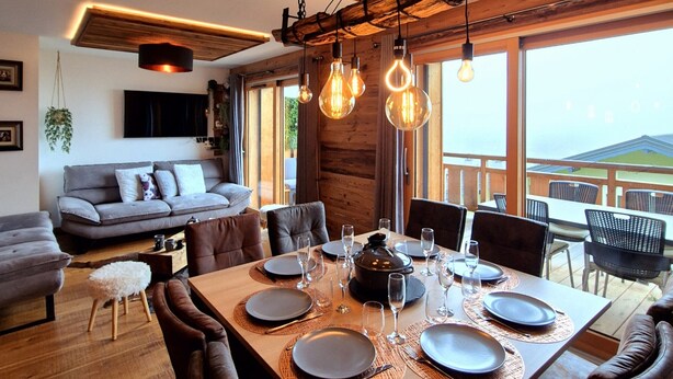 A dining area with a wooden table set for six, a sofa, and a TV mounted on the wall. - Cozy apartment in Les Gets, 3 bedrooms, Wi-Fi (Les Gets)