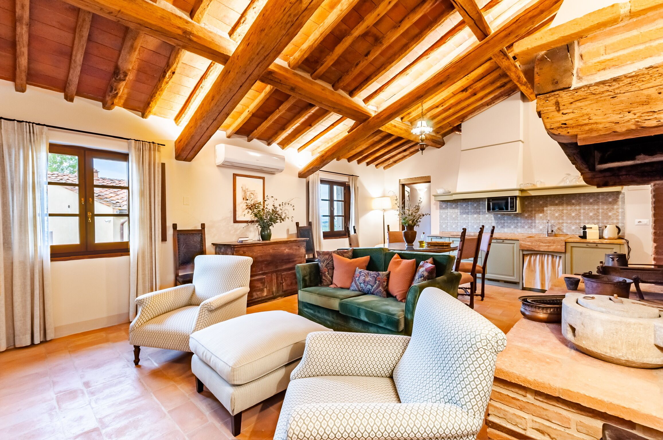 Ein geräumiges Wohnzimmer mit hoher Holzbalkendecke, einem grünen Sofa mit gemusterten Kissen sowie einer traditionellen Küchenzeile mit Holzschränken und einem Steinofen.