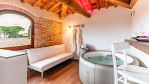 Une salle de spa avec un mur en pierre, des poutres en bois au plafond, un canapé blanc, un jacuzzi et une vue sur l’extérieur.