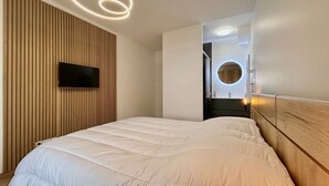 Une chambre moderne avec un grand lit, un téléviseur fixé sur un mur recouvert de panneaux en bois, et un miroir rond avec un éclairage intégré.