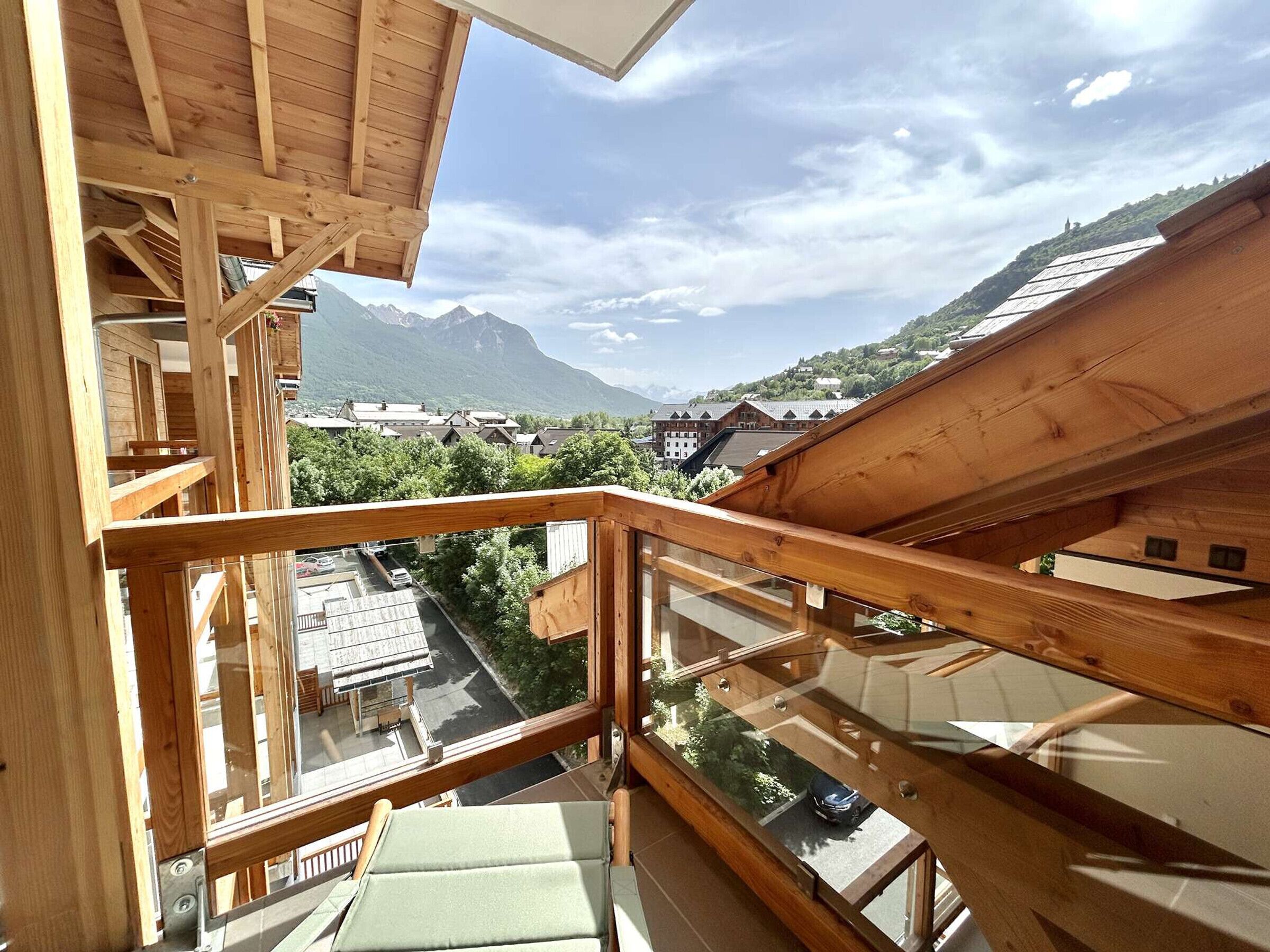 Un balcón de madera con barandilla de vidrio que da a un paisaje montañoso y un pueblo en la falda.