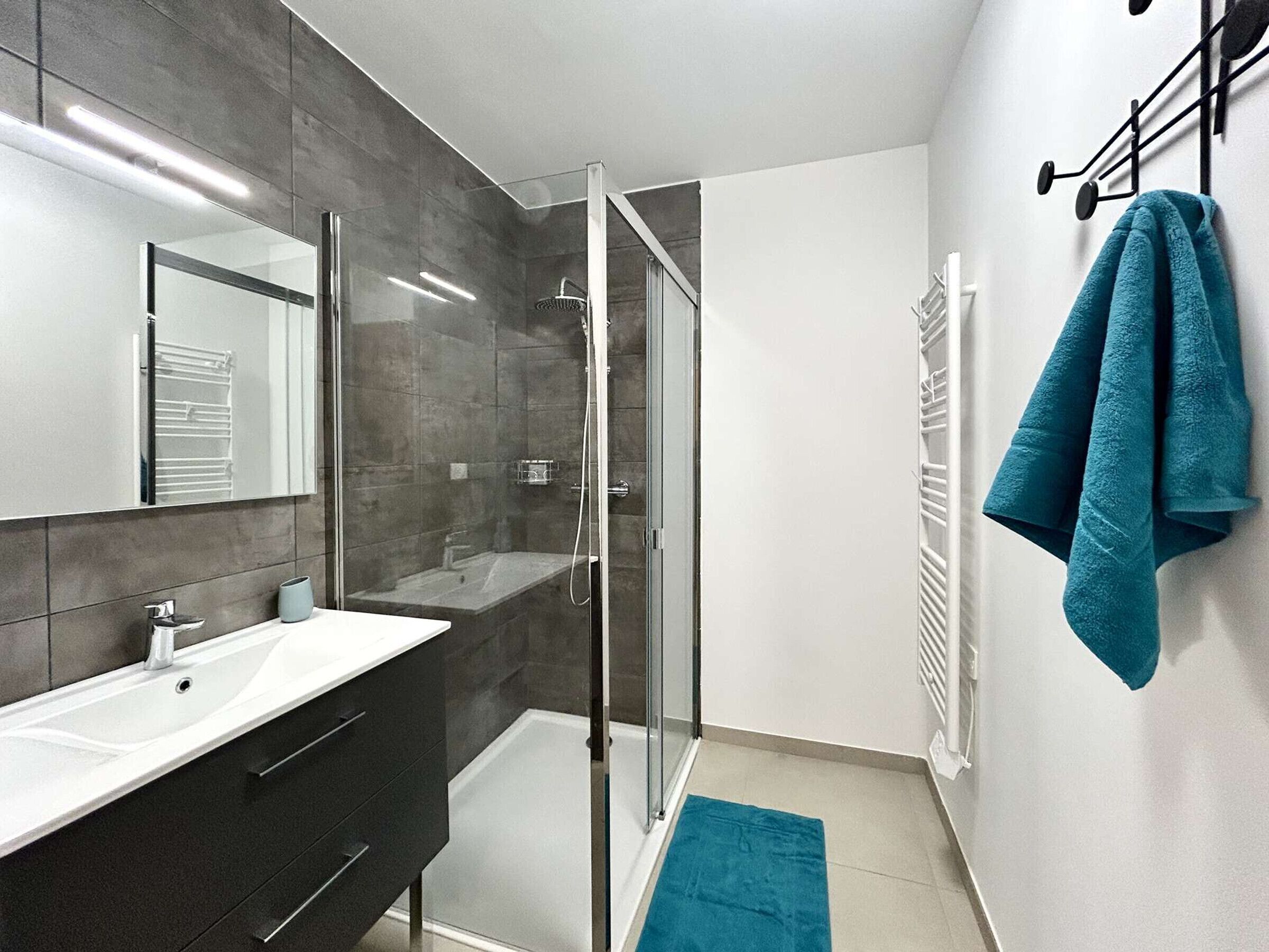 Un baño moderno con cabina de ducha de vidrio, un lavamanos blanco, muebles de color oscuro y una toalla verde azulado en un perchero.