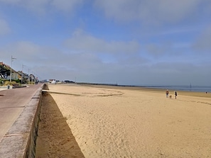 A beach with a paved walkway, a pier in the distance, and a clear sky.