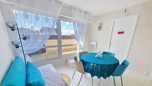 A room with a bed, a table with chairs, a window with curtains, and a door with a sign. - Cozy apartment 4 pax near sea with parking (Courseulles-sur-Mer)