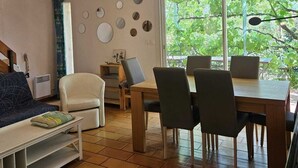Una sala de estar acogedora con un sofá, una mesita de centro y un comedor con sillas y una mesa. En la pared hay espejos decorativos circulares y una ventana con vistas a la vegetación.