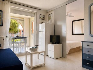 A compact hotel room with a bed, a desk with a computer, a small refrigerator, a TV, and a cart with cups.