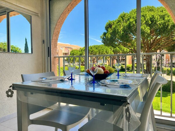 A glass-top table with a bowl of fruit and wine glasses, set against a backdrop of a clear blue sky and greenery.