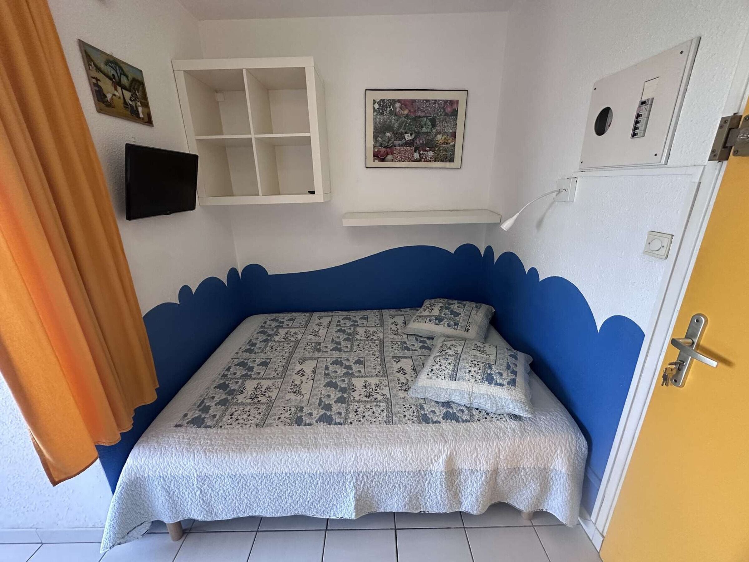 Une chambre avec un mur d’accent bleu, un lit avec une literie à motifs, un téléviseur fixé au mur et une petite étagère.
