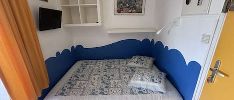 A bedroom with a blue accent wall, a bed with patterned bedding, a mounted TV, and a small shelf.