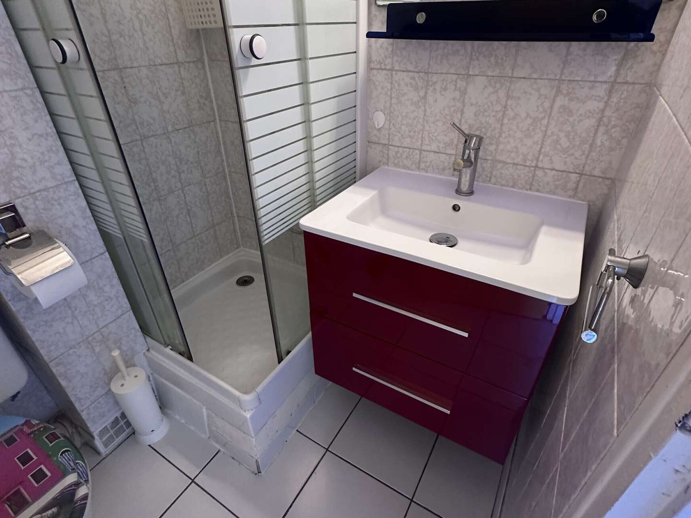 Ein Badezimmer mit Duschkabine aus Glas, einem weißen Waschbecken mit rotem Unterschrank und einer weißen Toilette.
