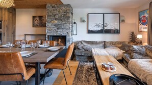 Un salon chaleureux avec une cheminée en pierre, un coin repas avec une table en bois dressée pour quatre personnes, et un espace salon confortable avec un canapé et des fauteuils.