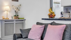Un salon avec un canapé gris, des coussins roses, une petite table avec une lampe et un panier de fruits.