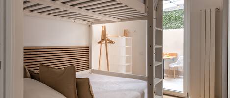 A bedroom with a bed, a wooden headboard, a white wall, a white ceiling, a white door, and a view of an outdoor area with a table and chairs.