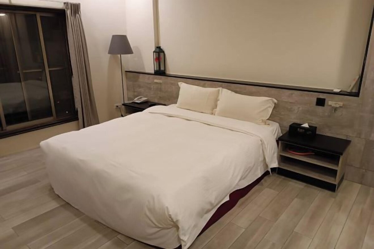 A hotel room with a large bed, a nightstand, a lamp, and a sliding door to the left.