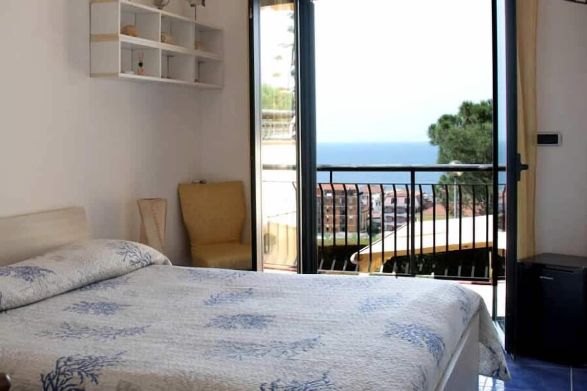 Un dormitorio con cama, una silla, estante y balcón con vista al mar.