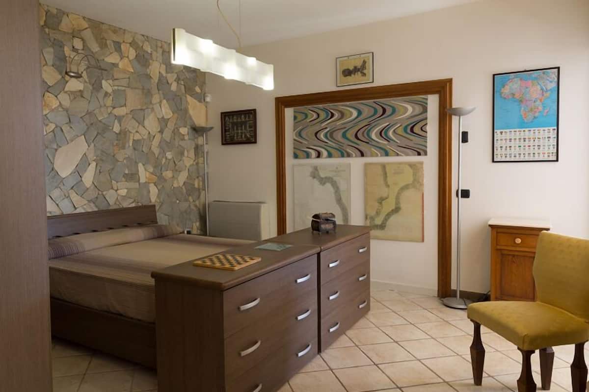 Un dormitorio con pared de piedra, una cama, un tocador, una silla y varias obras de arte en las paredes.