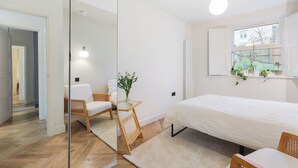 Un dormitorio moderno con una cama grande, una silla de madera, una mesita con un florero y una ventana con plantas.