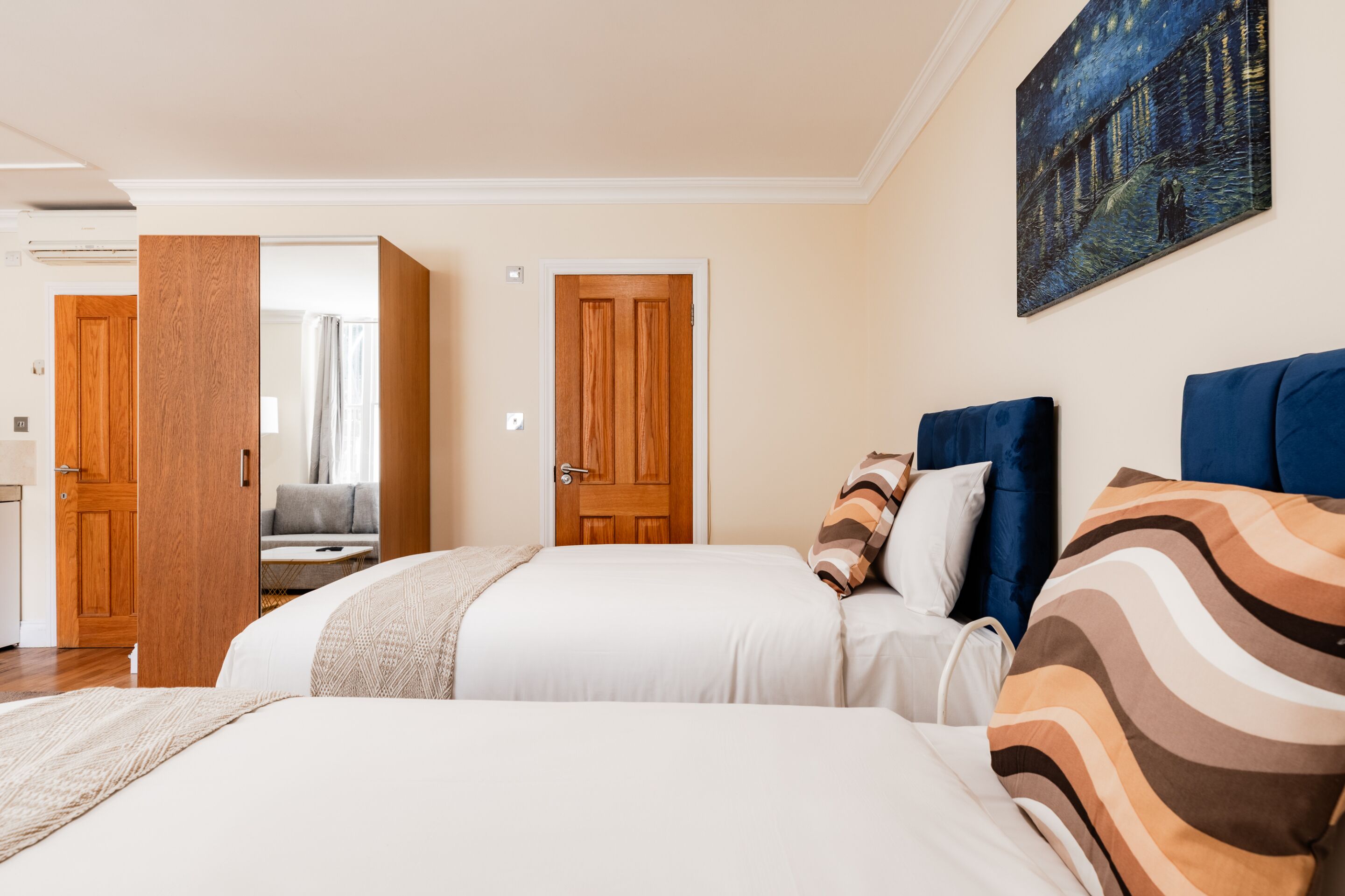 A hotel room with two beds, a wooden door, a painting on the wall, and a view of the hallway.