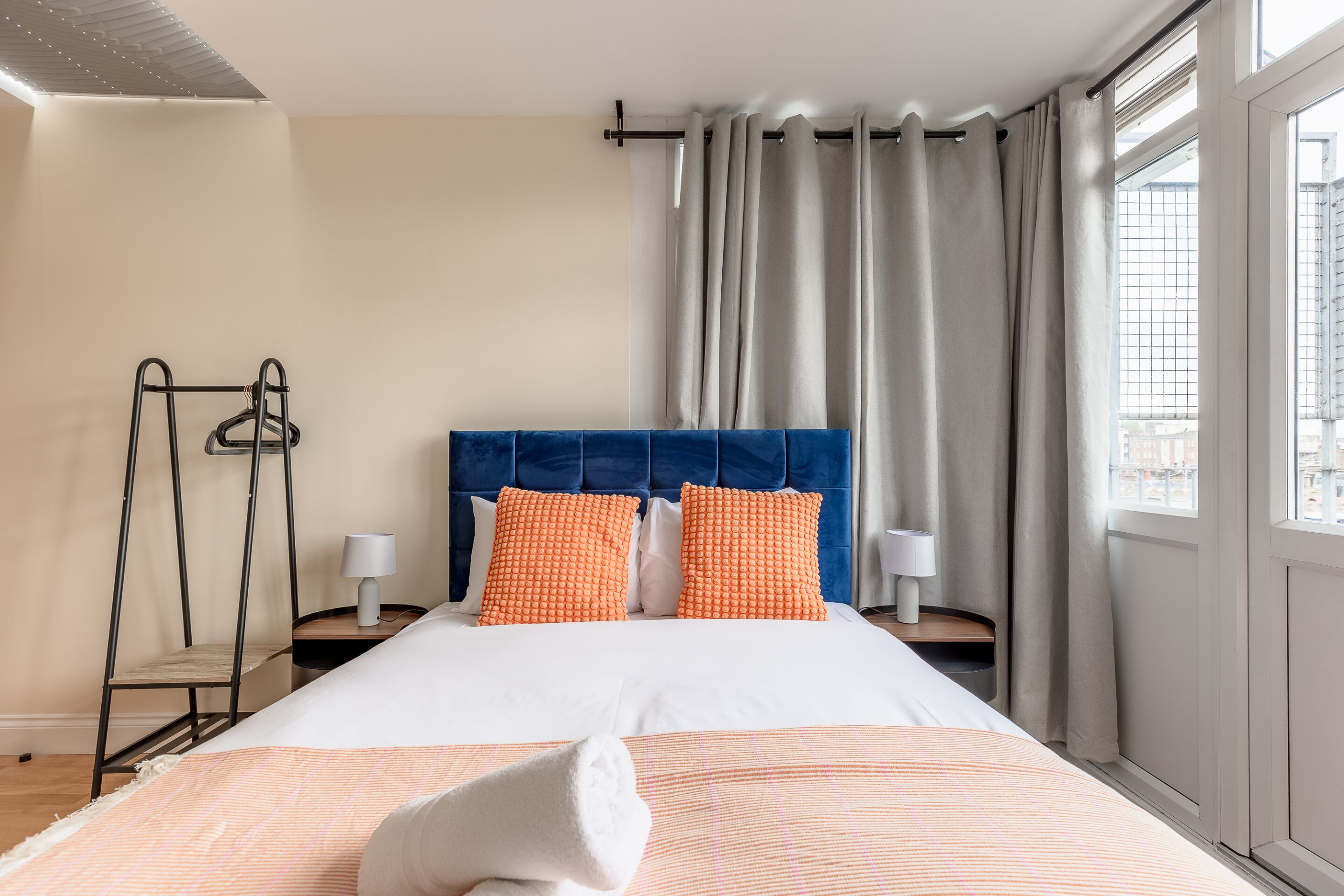 Ein Hotelzimmer mit einem Bett, zwei Kissen, einem Nachttisch, einer Lampe, einem Hängeregál und einem Fenster mit Vorhängen.