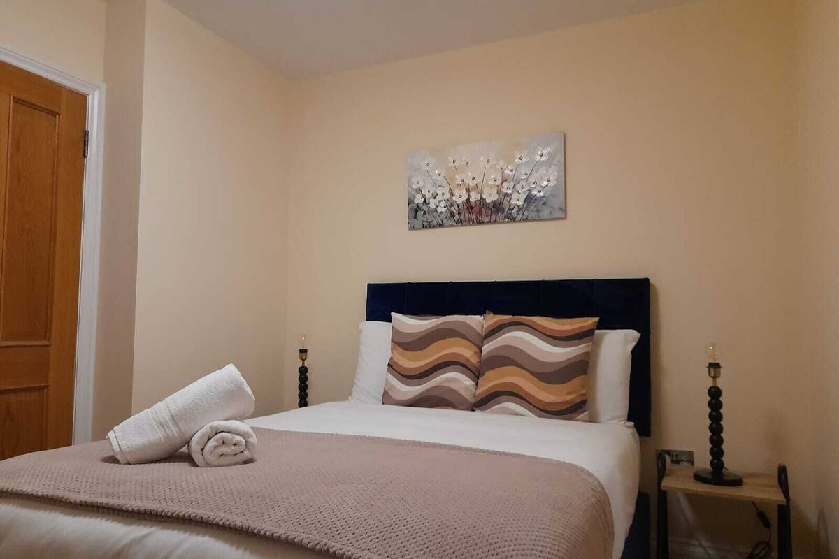 A neatly made bed with a beige blanket and pillows, a wall-mounted artwork, and a bedside table with a lamp.